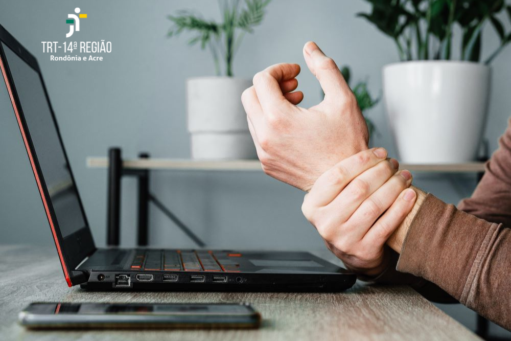 A imagem mostra um notebook e um celular sobre a mesa e as mãos de uma pessoa fazendo referência a dor.