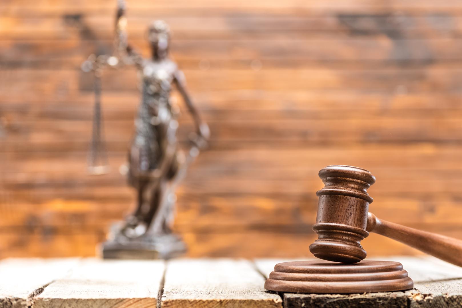 Vista de perto do martelo de madeira na mesa e ao fundo a estatueta de Themis, a deusa da justiça.